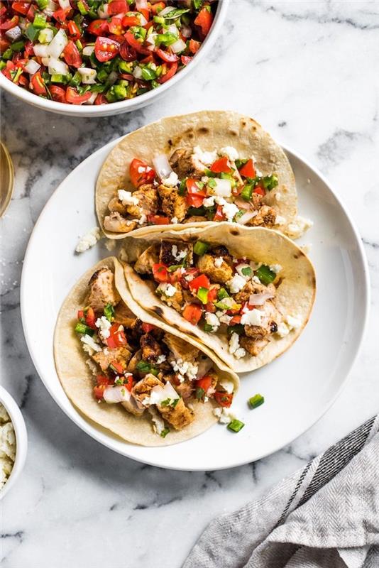 tortillainpackningar, fyllda med grönsaker, kycklingfilé, arrangerade på vit tallrik, taco med hårt skal, bänkskiva i marmor