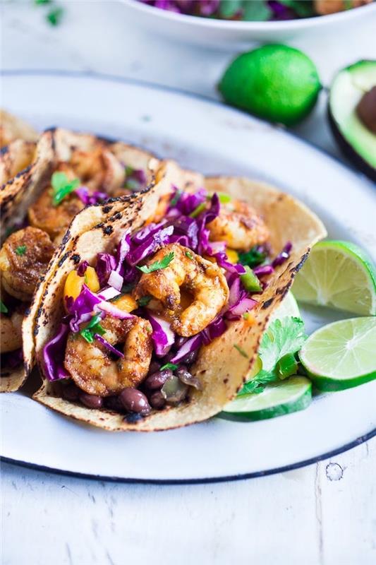 tortillainpackningar, fyllda med kål, stekta räkor, på en vit tallrik, tacoingredienser, limeskivor på sidan